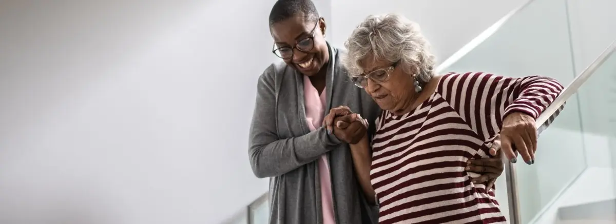 Female caregiver helping senior woman walk down stairs.