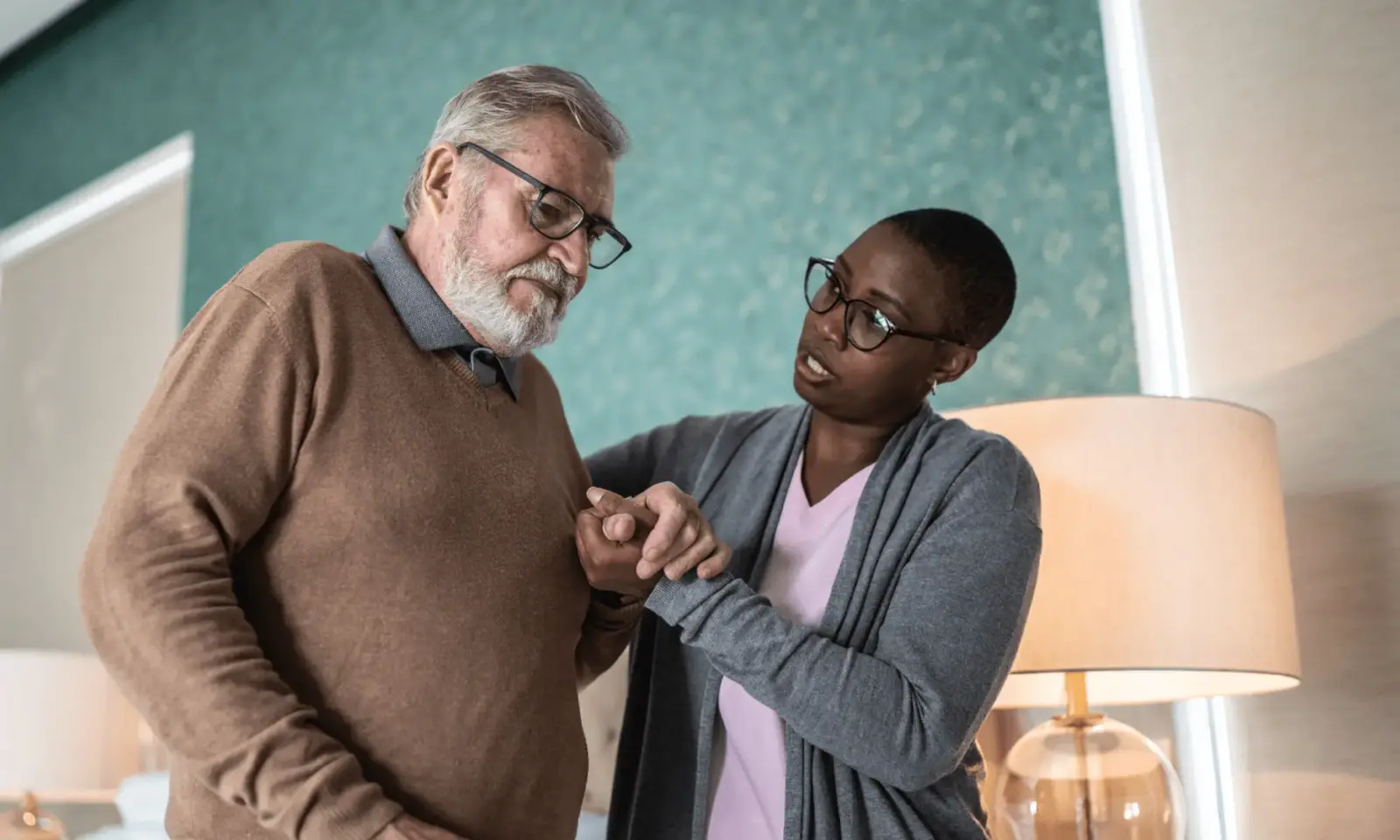 Caregiver wearing a pink shirt assists an older gentleman dealing with Parkinson’s Disease in his home.