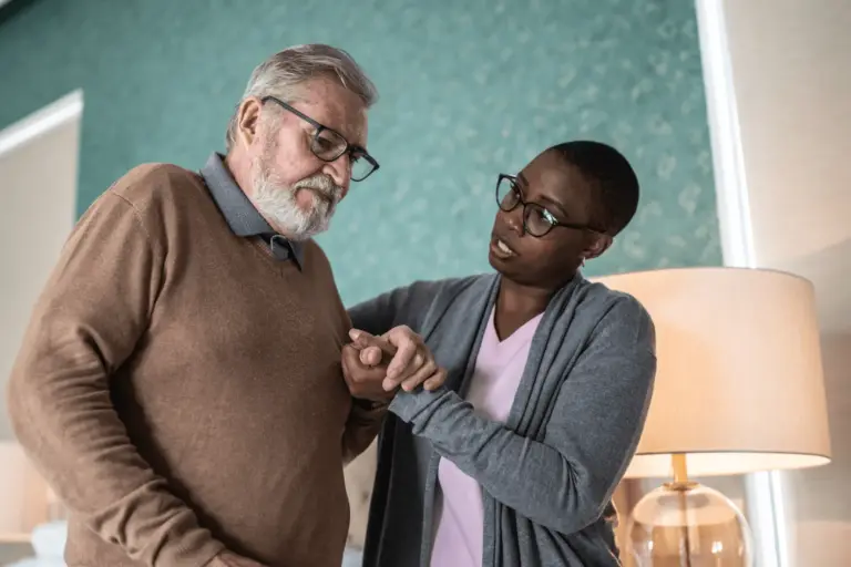 Caregiver wearing a pink shirt assists an older gentleman dealing with Parkinson’s Disease in his home.