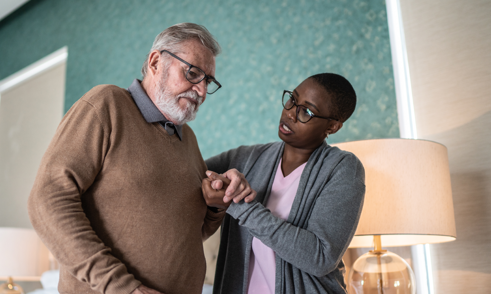 Caregiver wearing a pink shirt assists an older gentleman dealing with Parkinson’s Disease in his home.