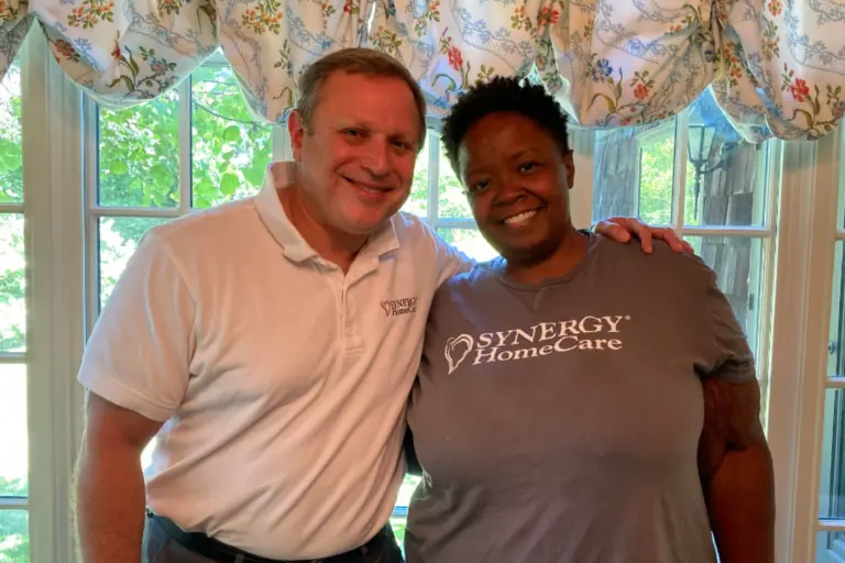 SYNERGY HomeCare franchise owner, Ron Schulman is wearing a white SYNERGY HomeCare shirt next to stands next to Mary, wearing a gray SYNERGY HomeCare shirt.