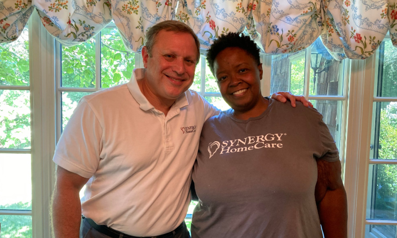 SYNERGY HomeCare franchise owner, Ron Schulman is wearing a white SYNERGY HomeCare shirt next to stands next to Mary, wearing a gray SYNERGY HomeCare shirt.