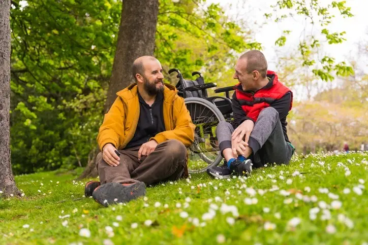 gentleman in an orange-colored jacket talks to a man living with a disability on the grass at a local park.