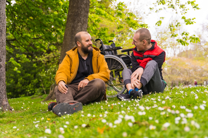 gentleman in an orange-colored jacket talks to a man living with a disability on the grass at a local park.