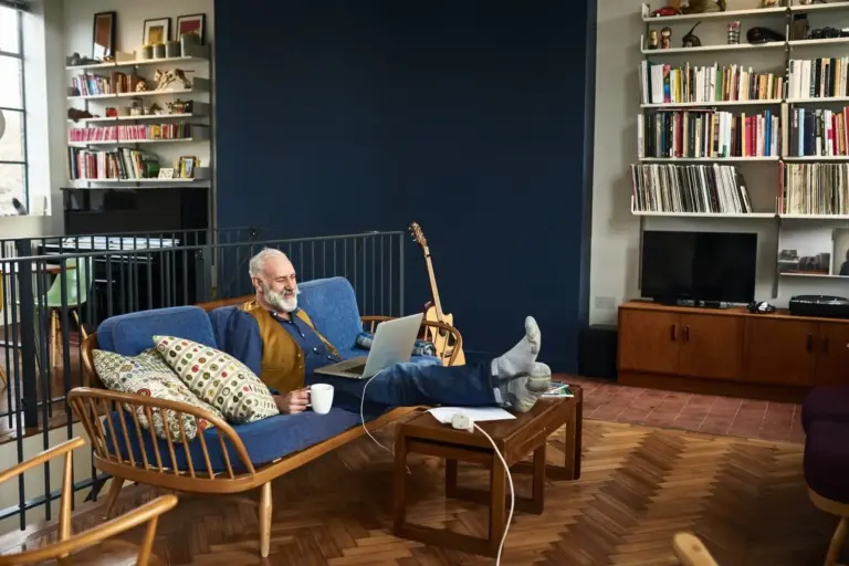 An older man sits with a laptop on his lap and a cup of coffee while speaking to a long distance family caregiver.