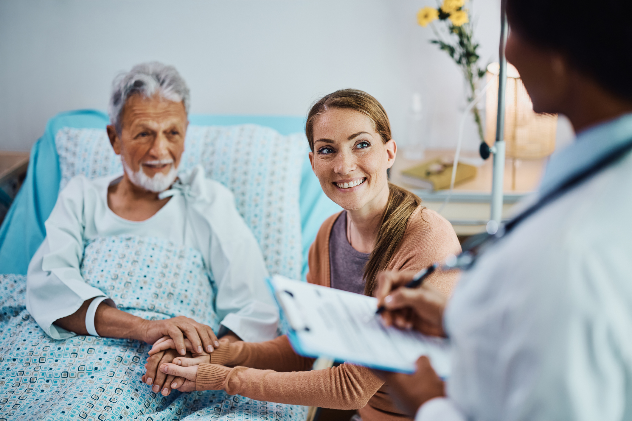 Male senior patient in hospital bed and young woman sitting next to him smile toward doctor