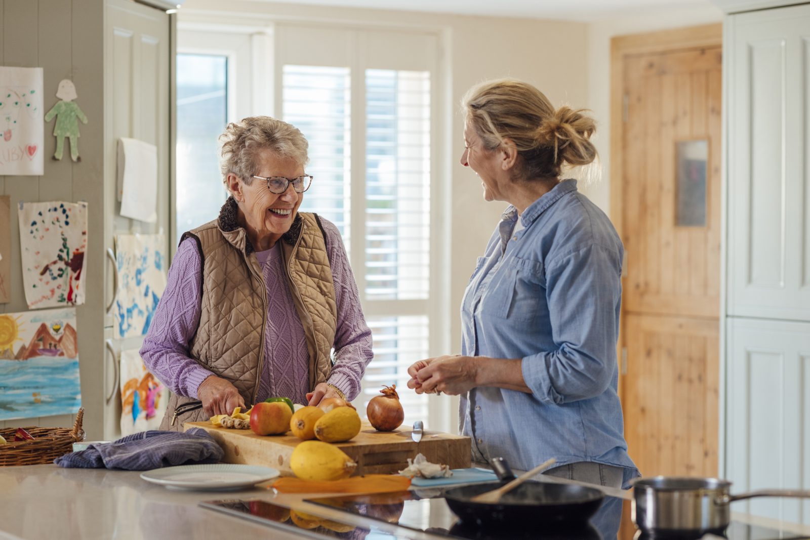 A senior woman and her in-home caregiver laugh and chop vegetables in the senior woman’s kitchen as they both cook a healthy meal together.