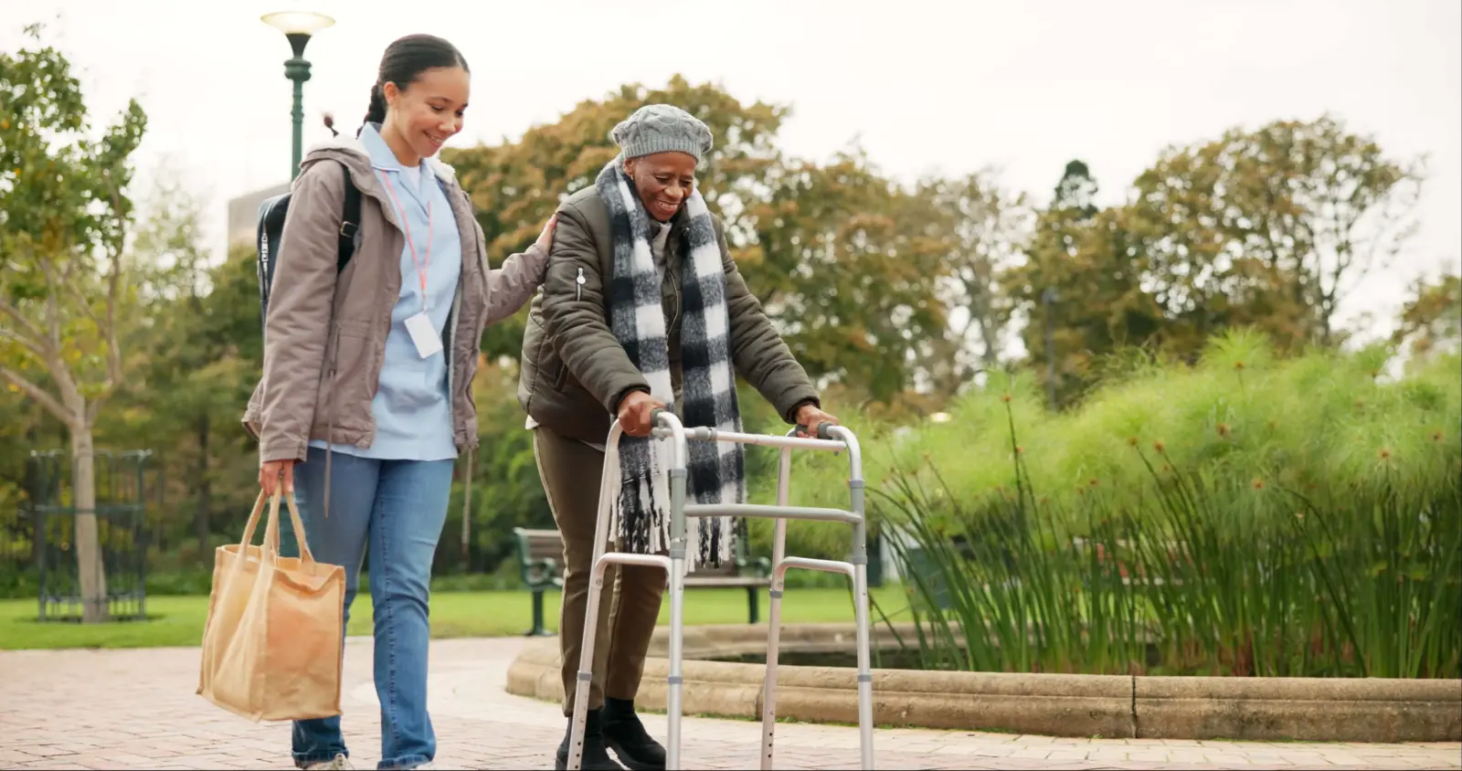 Friendly caregiver provides companionship and support as she walks outside in a park alongside a smiling senior woman using a walker.