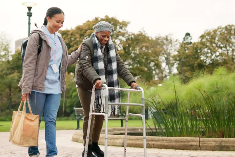 Friendly caregiver provides companionship and support as she walks outside in a park alongside a smiling senior woman using a walker.