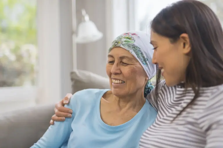 Senior woman with cancer is smiling while wearing a headscarf and receiving a side hug from her smiling daughter.
