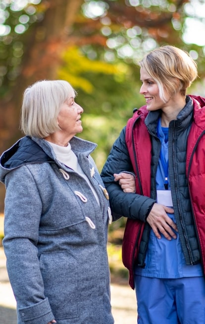 Female caregiver walking along side a senior woman with hand in arm to support her and prevent falls.