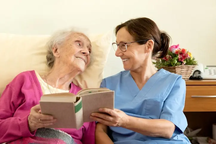 An adult daughter who is a paid caregiver for her mother reads a book out loud with her aging mother in bed.