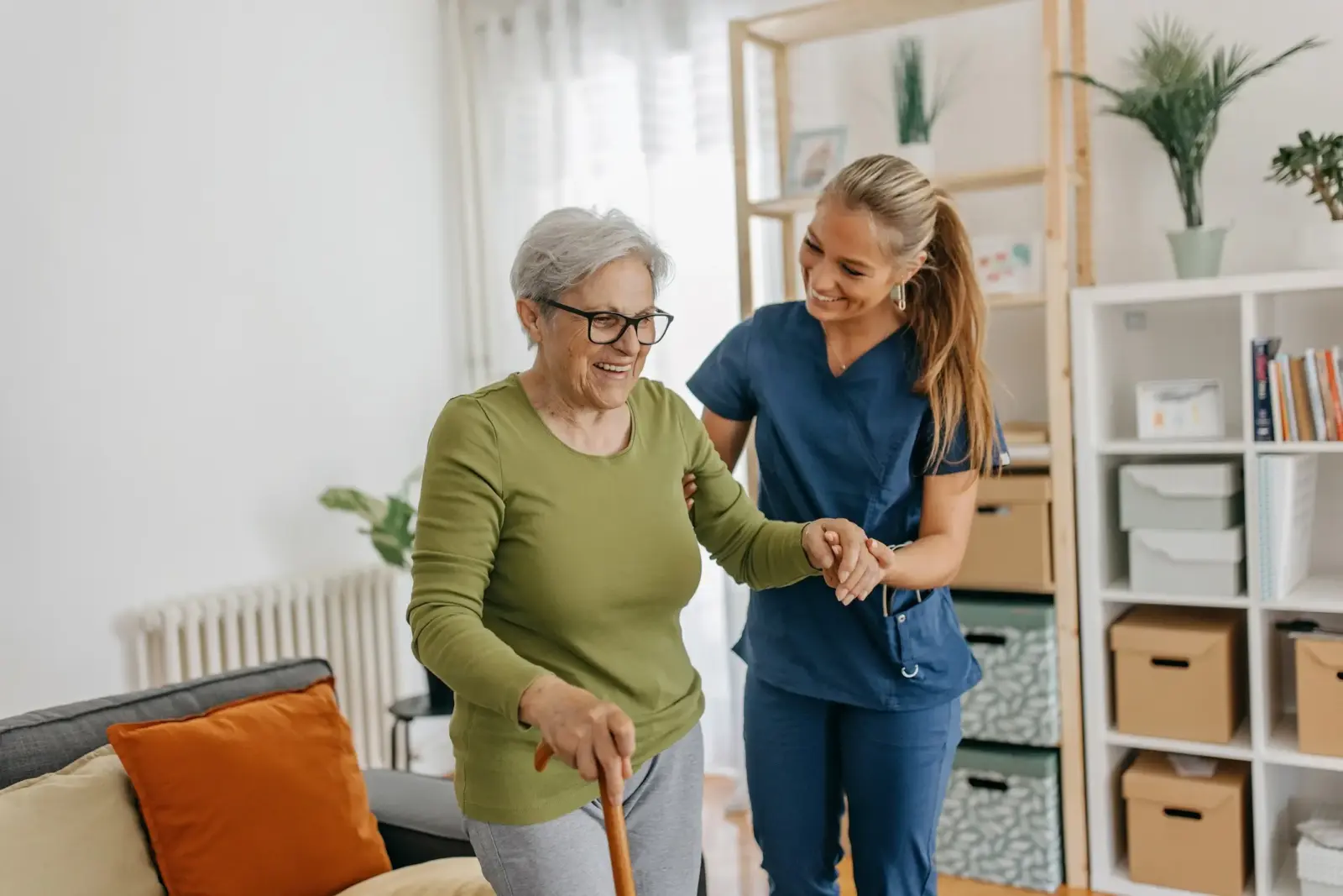 Smiling female caregiver provides light physical assistance for happy senior woman walking through her living room with a cane.