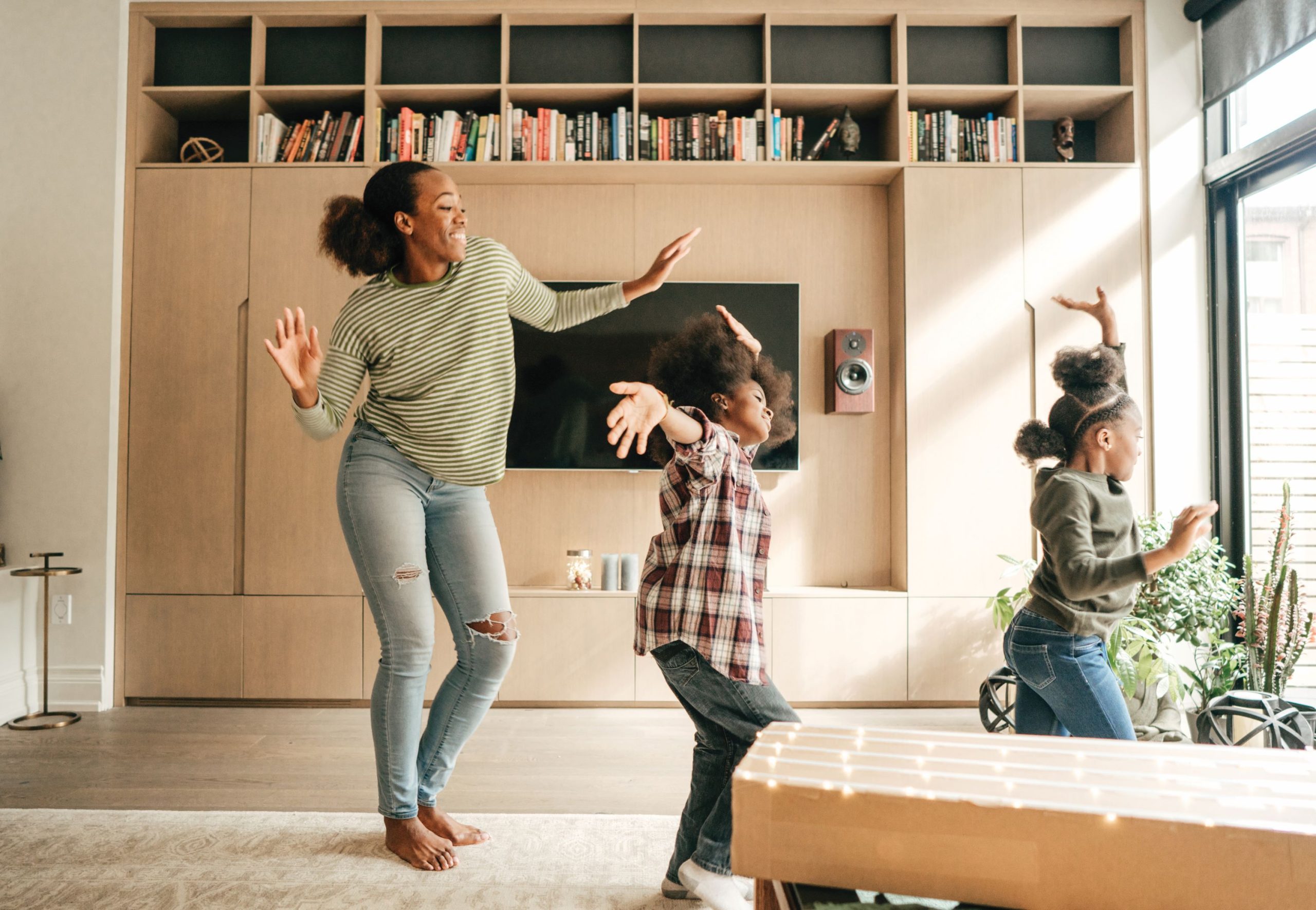 Young woman and two children enjoy back up child care while dancing around a living room.
