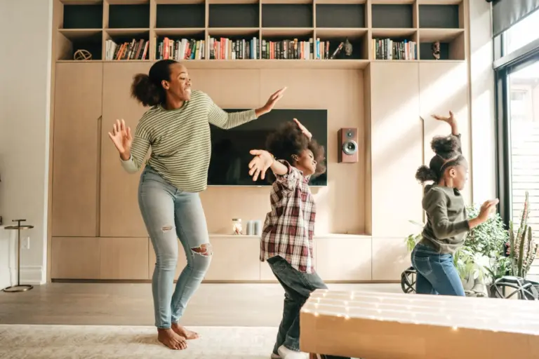Young woman and two children enjoy back up child care while dancing around a living room.