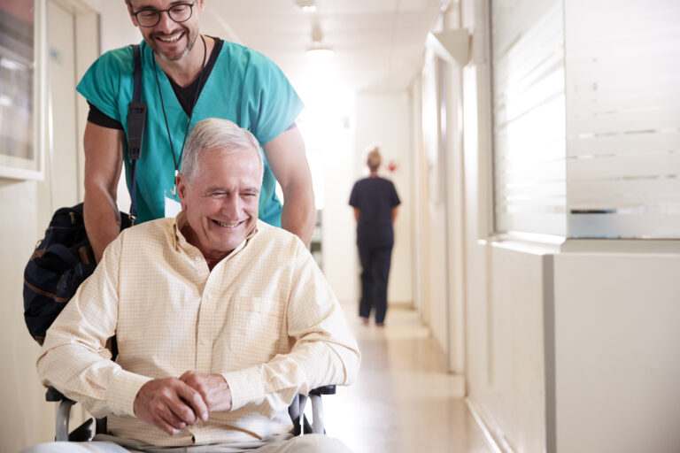 Male Orderly Pushing Senior Male Patient Being Discharged From Hospital In Wheelchair