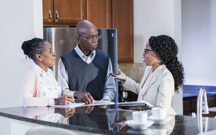 A senior African-American couple in their 70s at home in the kitchen talking with a financial advisor. The consultant is a mature African-American woman in her 40s.