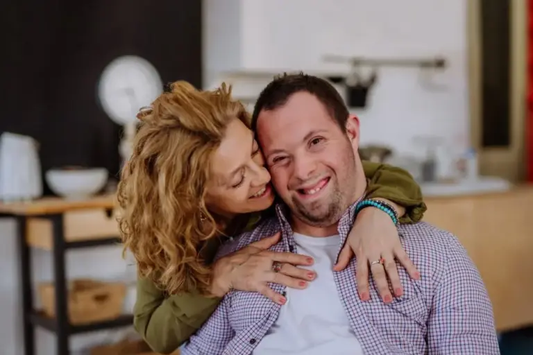 A disabled gentleman sits down while his partner hugs him.