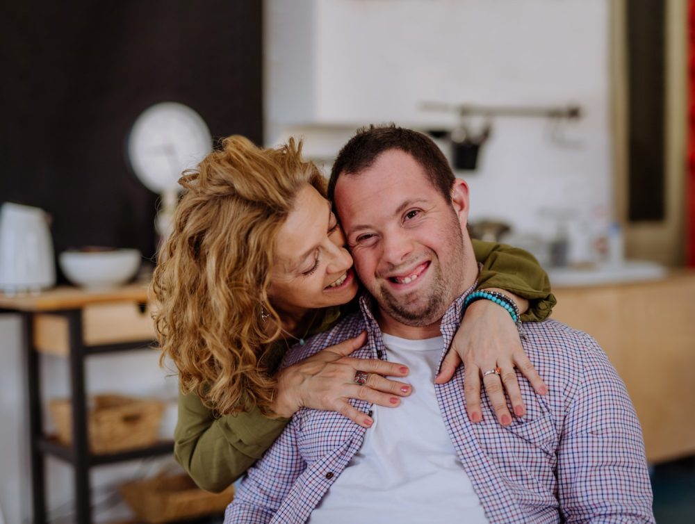 A disabled gentleman sits down while his partner hugs him.