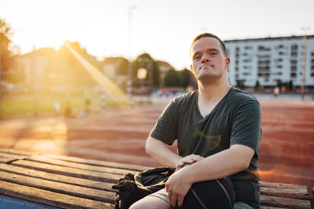 A gentleman living with a developmental disability is sitting on a bench by a track during sunrise.
