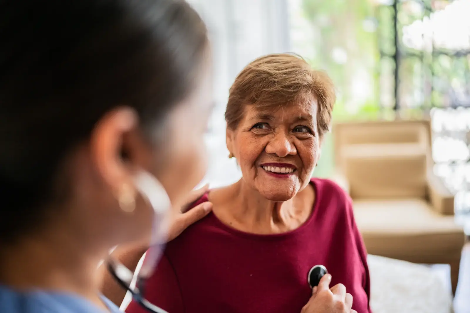 Home caregiver listening to a smiling senior woman's heartbeat in her home.