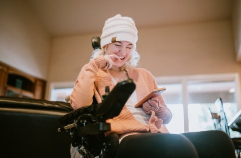 Young woman with physical disability seated in her wheelchair looking at a phone and laughing.