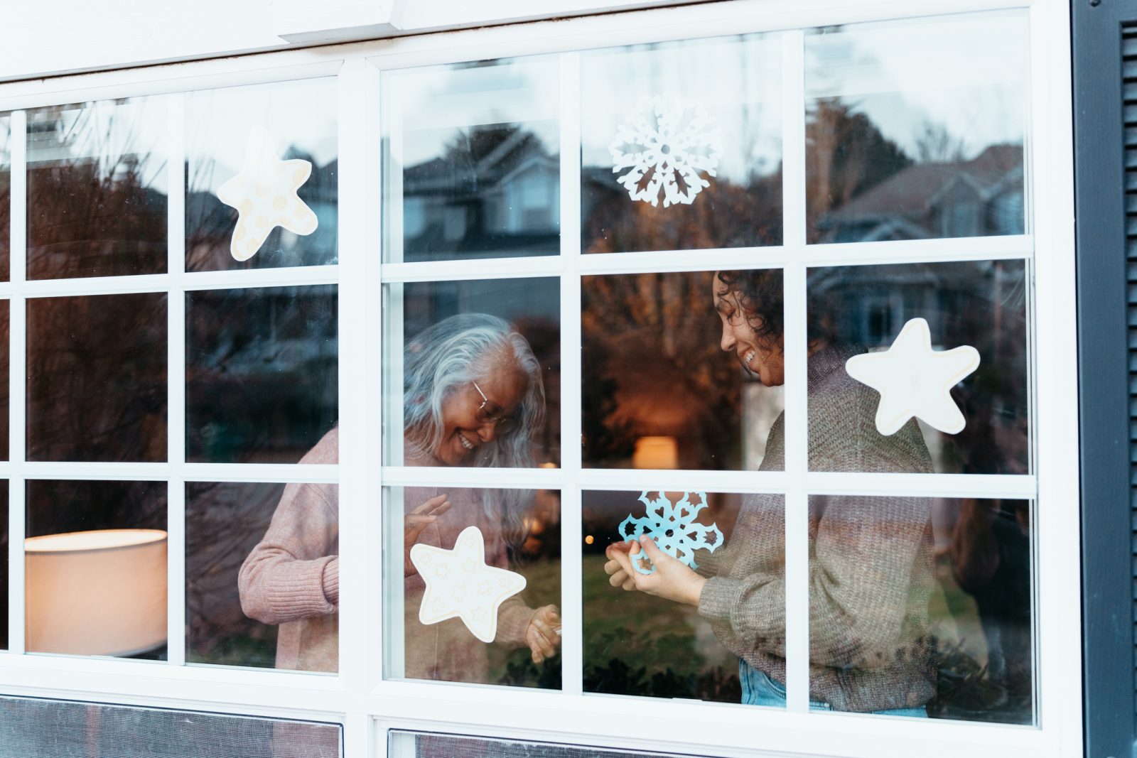 Senior woman and adult daughter at home hanging snowflakes in window