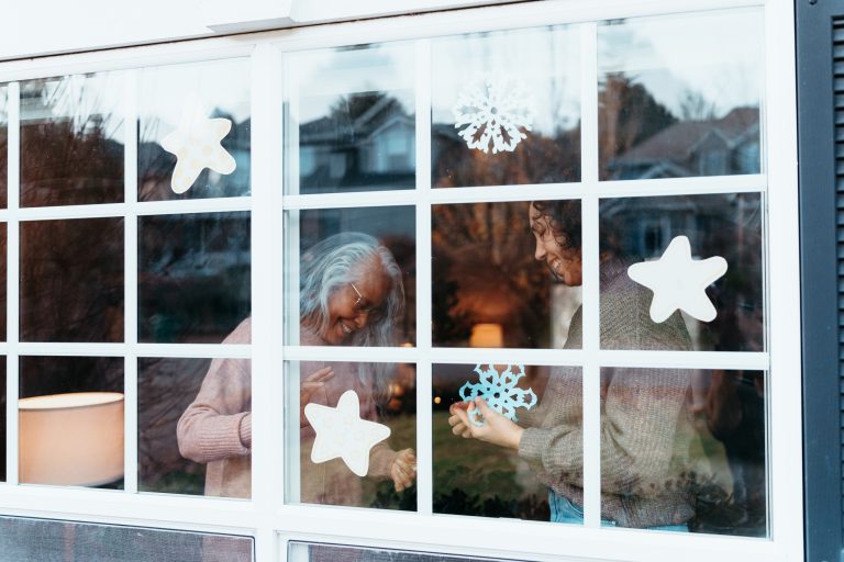 Senior woman and adult daughter at home hanging snowflakes in window