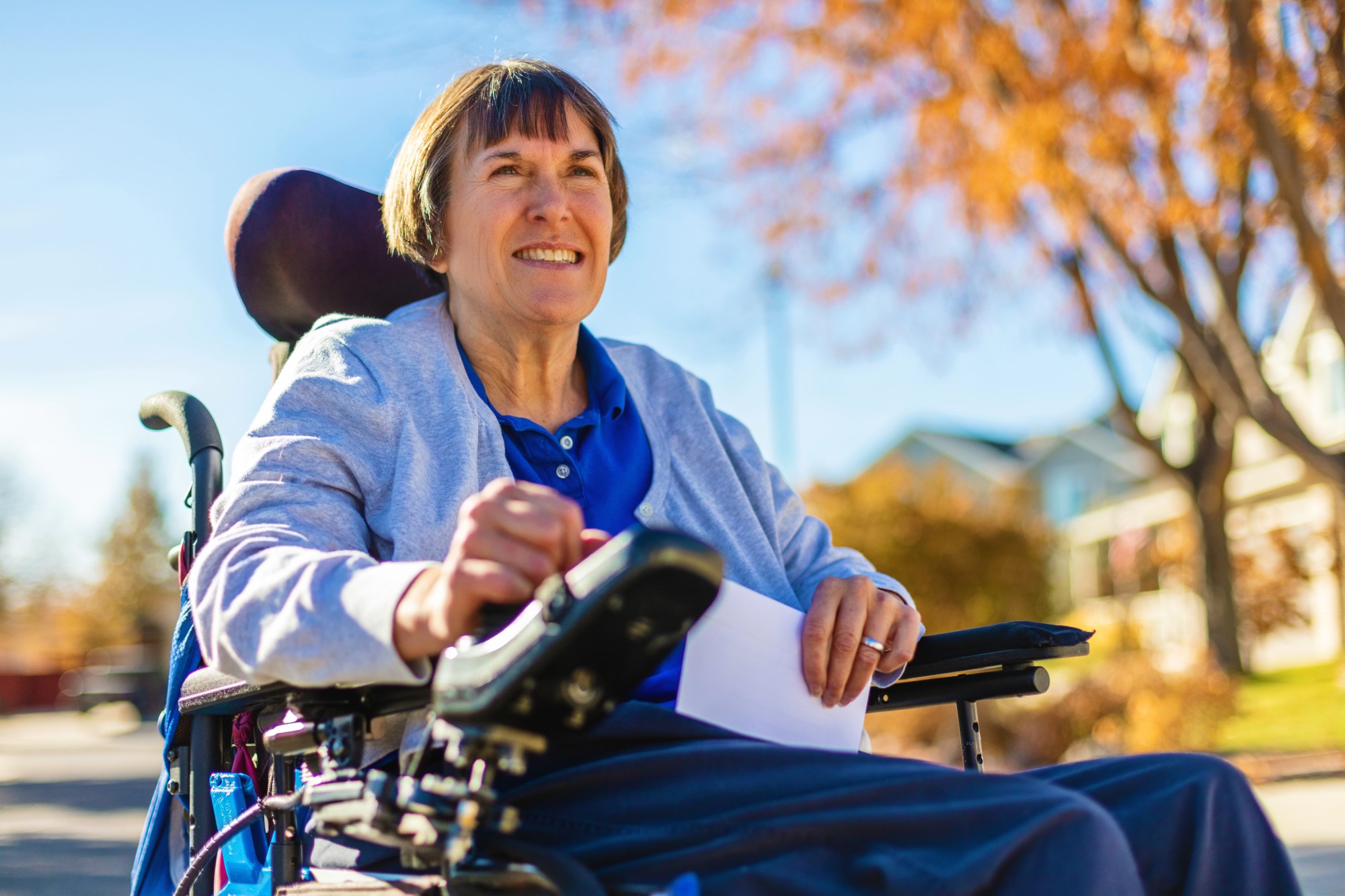 A woman wearing a blue button-up shirt living with a disability navigates down her street to drop mail off at a nearby mailbox with her power chair.