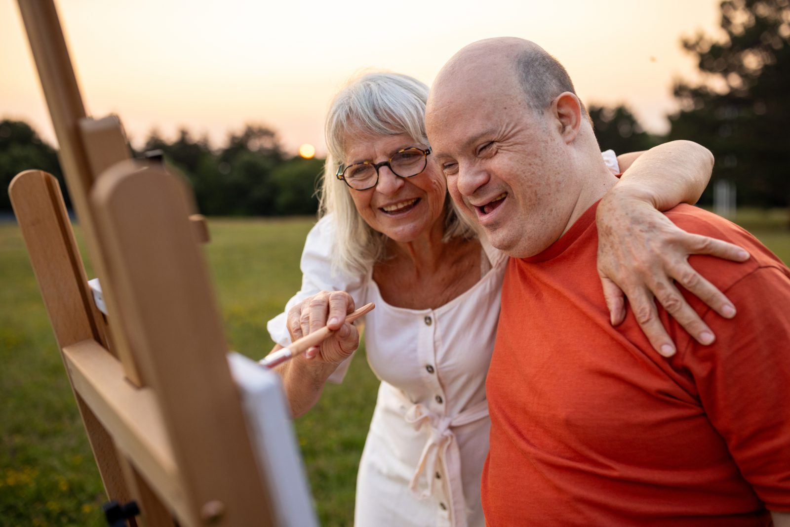 Two people, man with down syndrome and woman artist painting together outdoors on a meadow.