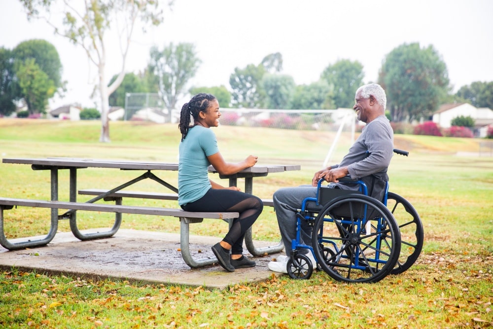 SYNERGY HomeCare caregiver sitting with senior man in wheelchair at park.