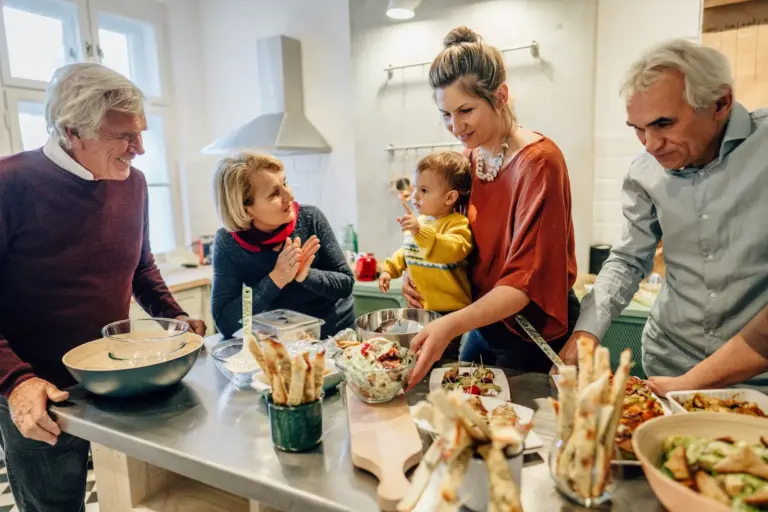 Middle-aged woman, who is a Sandwich Generation caregiver, prepares a meal in her kitchen alongside her child and aging loved ones.