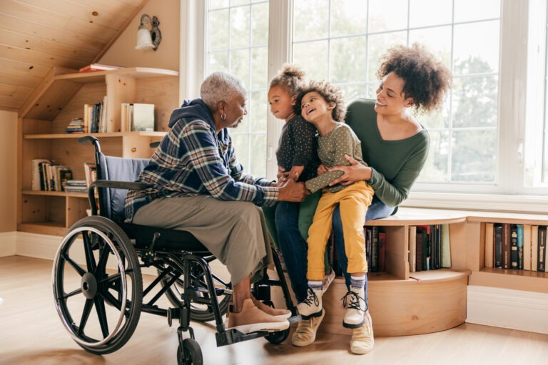 A smiling young woman holds her two children as they cheerfully visit their grandmother who is in a wheelchair.