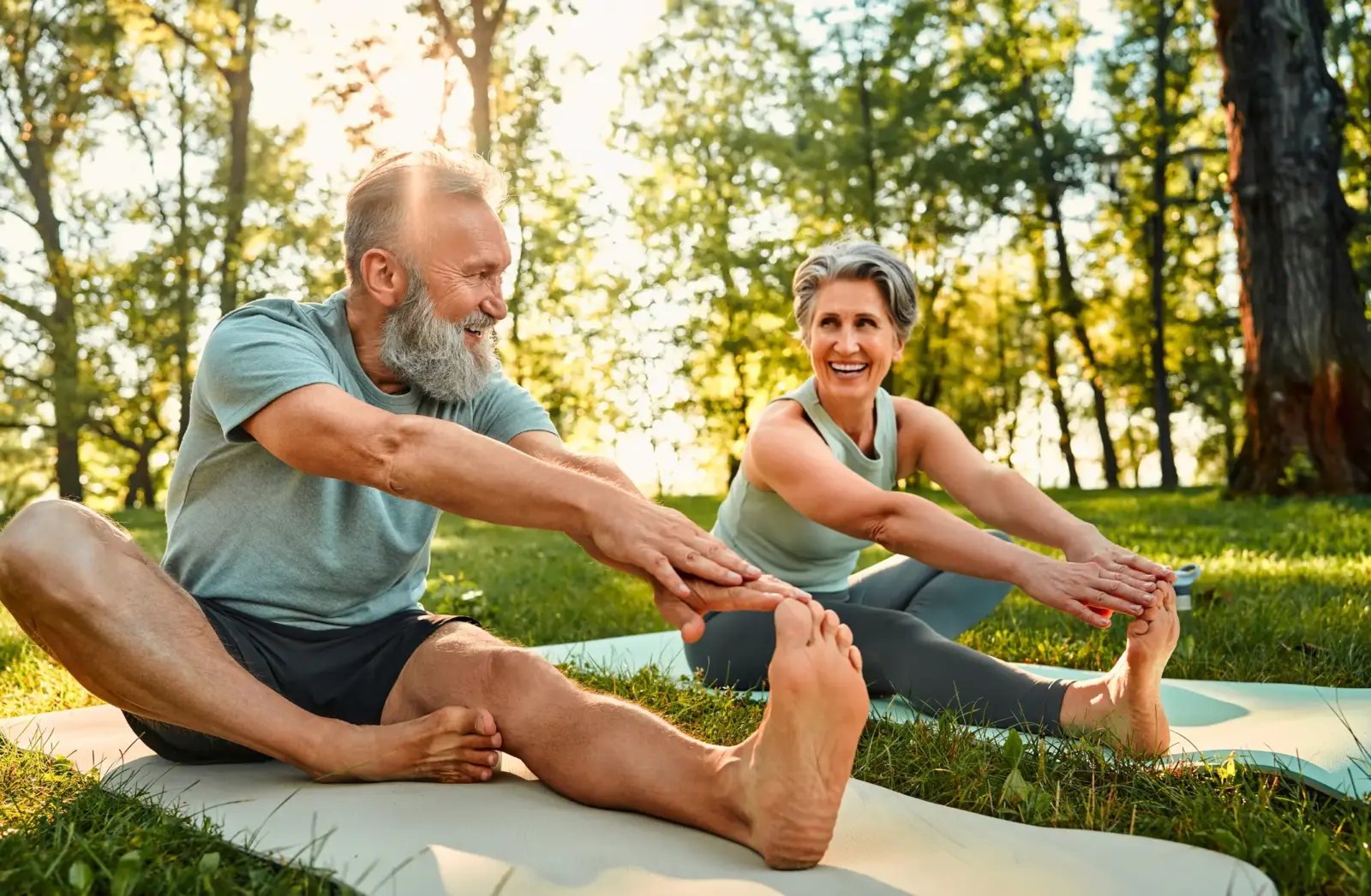 Happy senior couple stretching on yoga mats with hands to one leg during outdoors workout in sunny park.