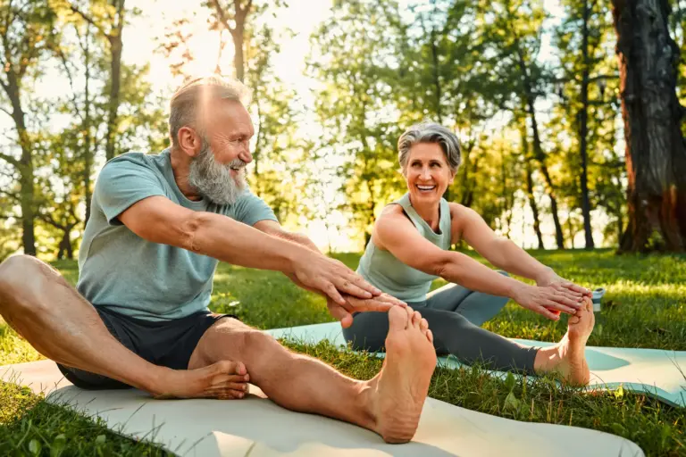 Happy senior couple stretching on yoga mats with hands to one leg during outdoors workout in sunny park.