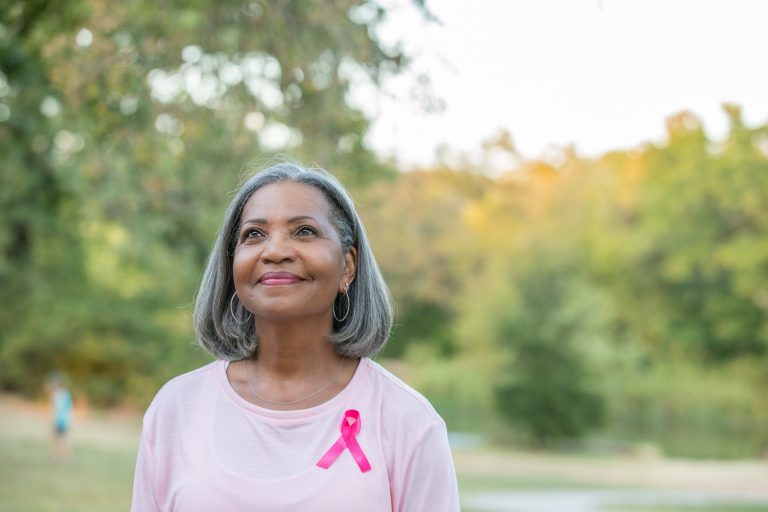 Senior woman smiles while walking outside for breast cancer awareness and wearing a pink breast cancer ribbon pinned to her shirt