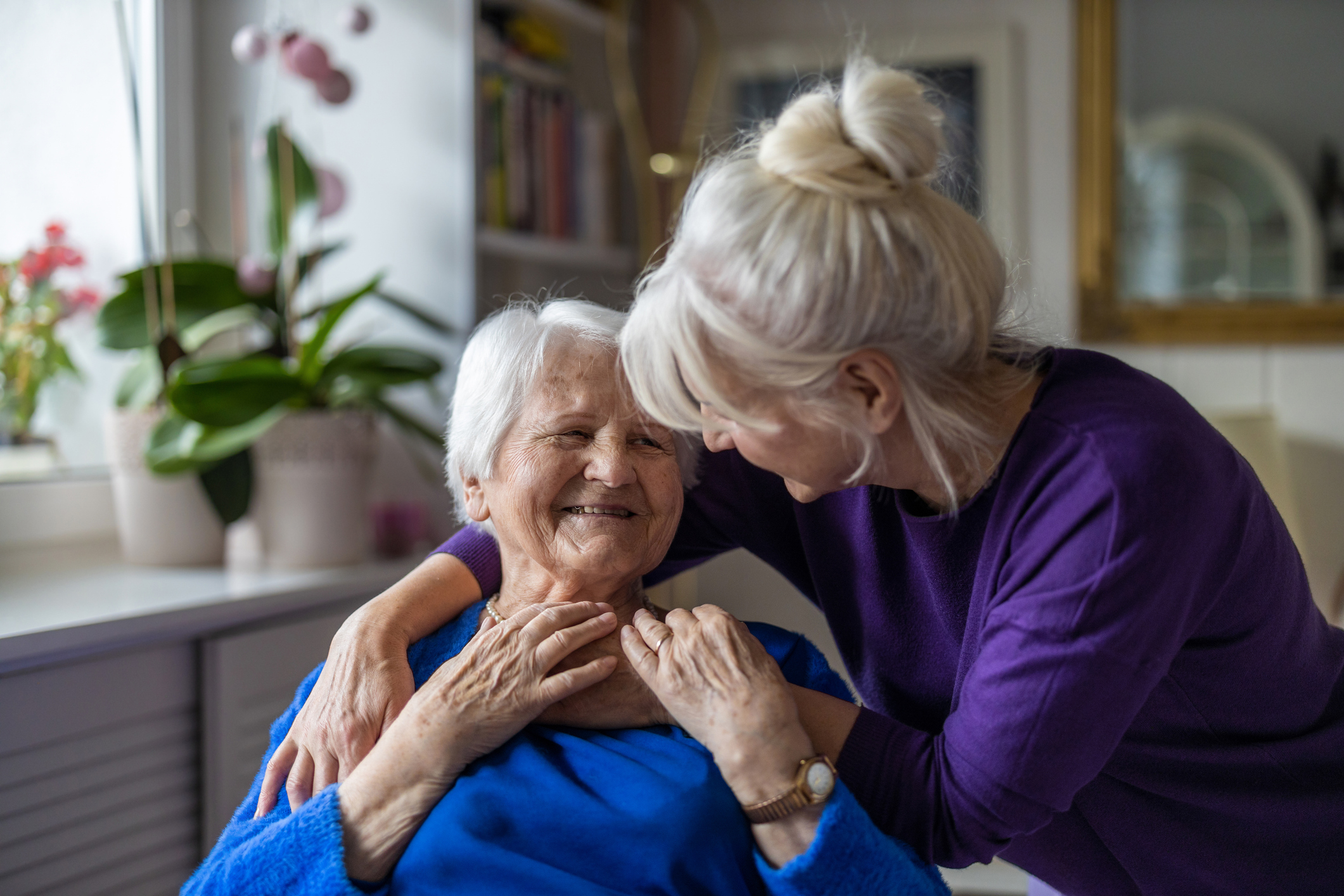 Senior woman smiling in living room, embraced by adult daughter, symbolizing support during Alzheimer’s & Brain Awareness Month.