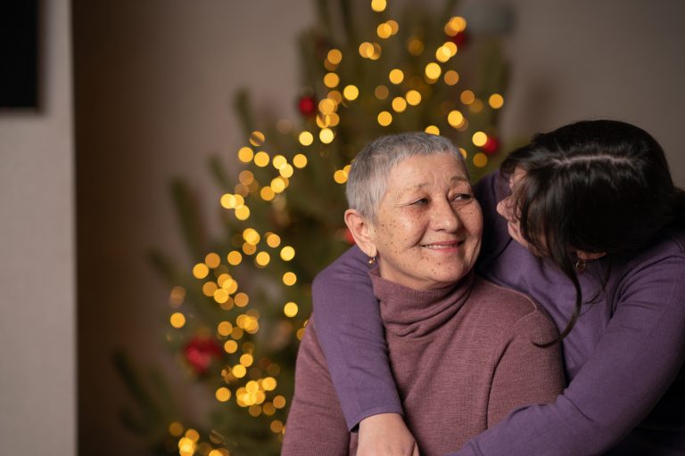 Senior woman smiles at daughter who is giving her a hug in front of their holiday decorations.