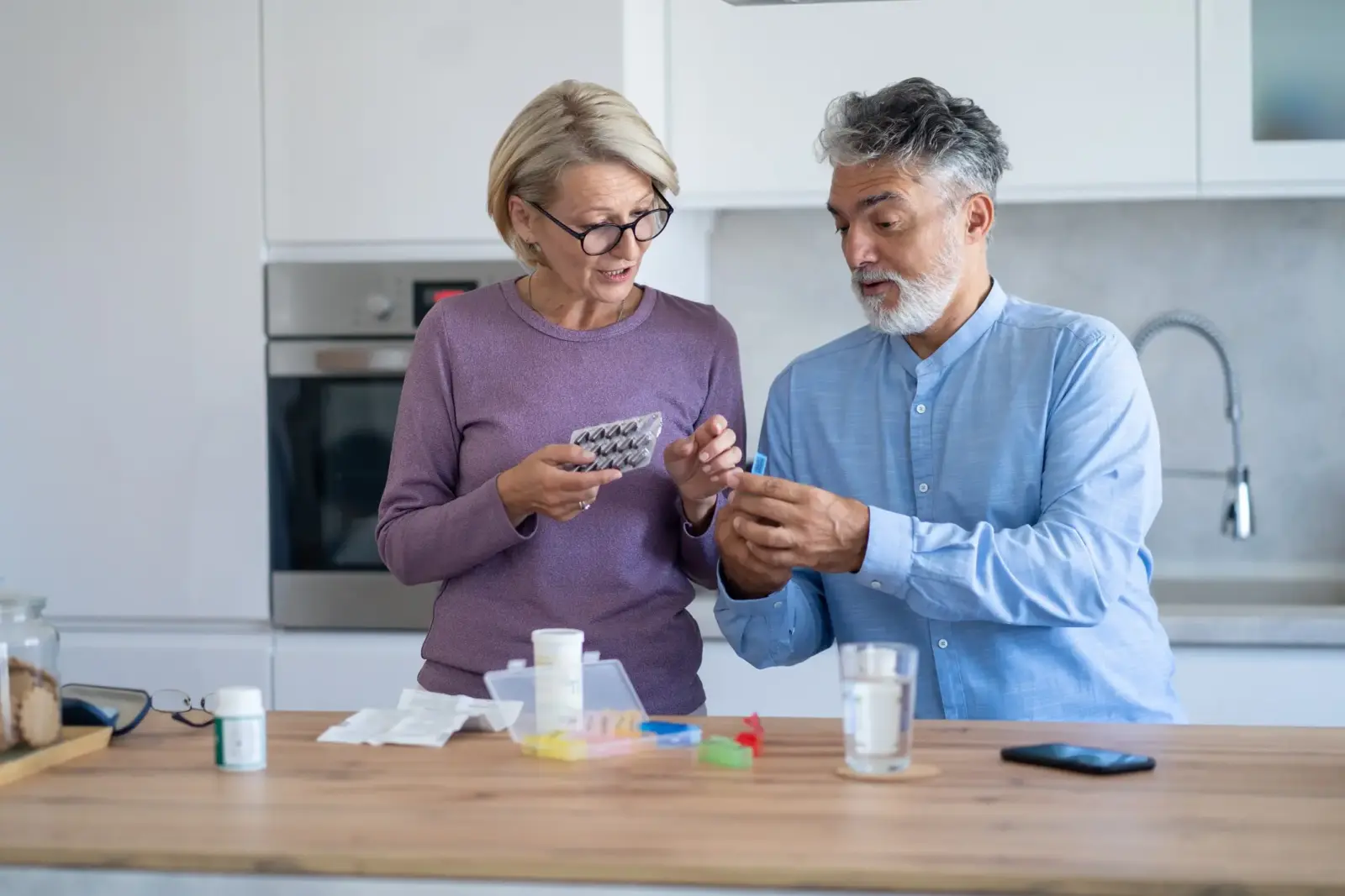 A mature gray-haired married couple helping each other organize their daily medication into a pill organizer in their kitchen