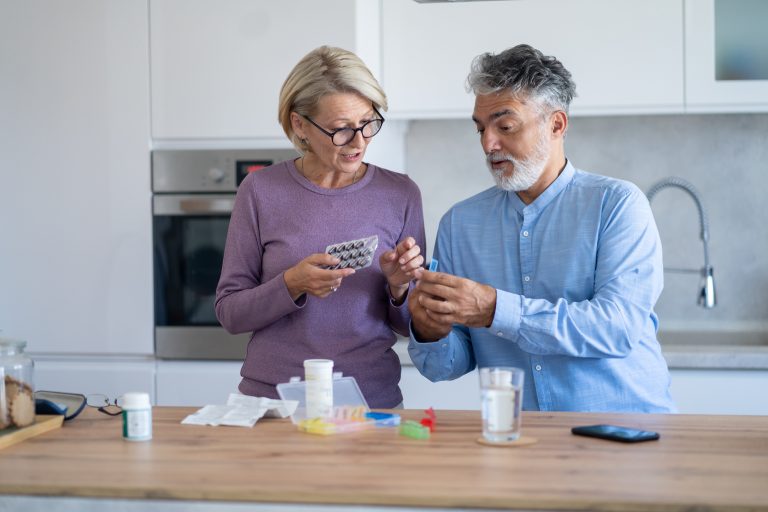 A mature gray-haired married couple helping each other organize their daily medication into a pill organizer in their kitchen