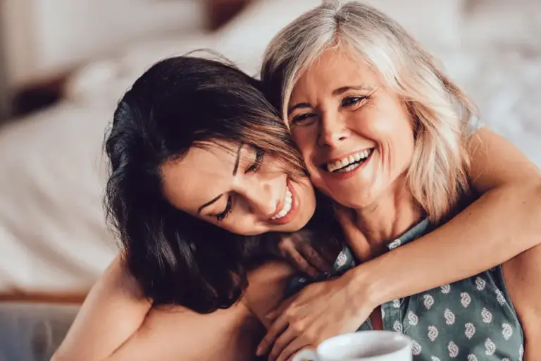 A woman hugs her mother who is sitting and smiling.