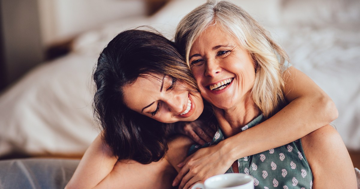 A woman hugs her mother who is sitting and smiling.