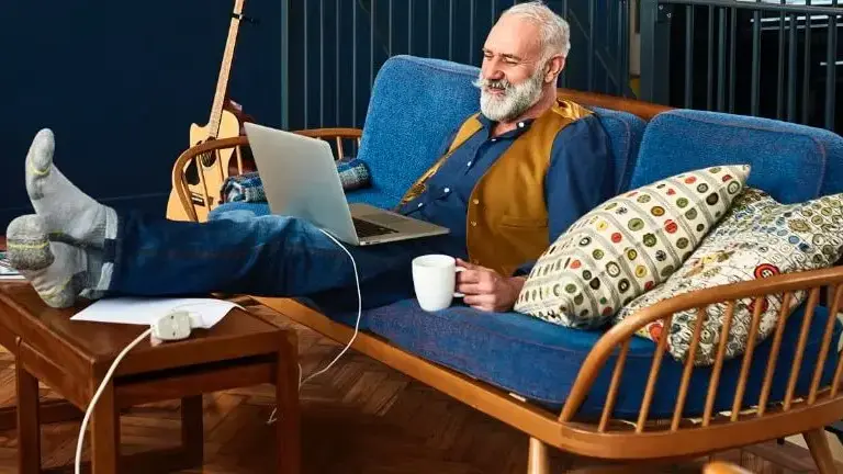 Senior man sitting on the couch looking up the cost of home care near him on his laptop.