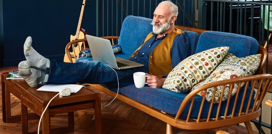 Senior man sitting on the couch looking up the cost of home care near him on his laptop.