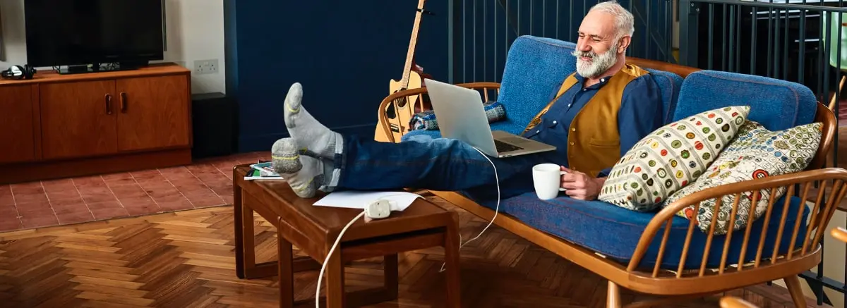 Senior man sitting on the couch looking up the cost of home care near him on his laptop.