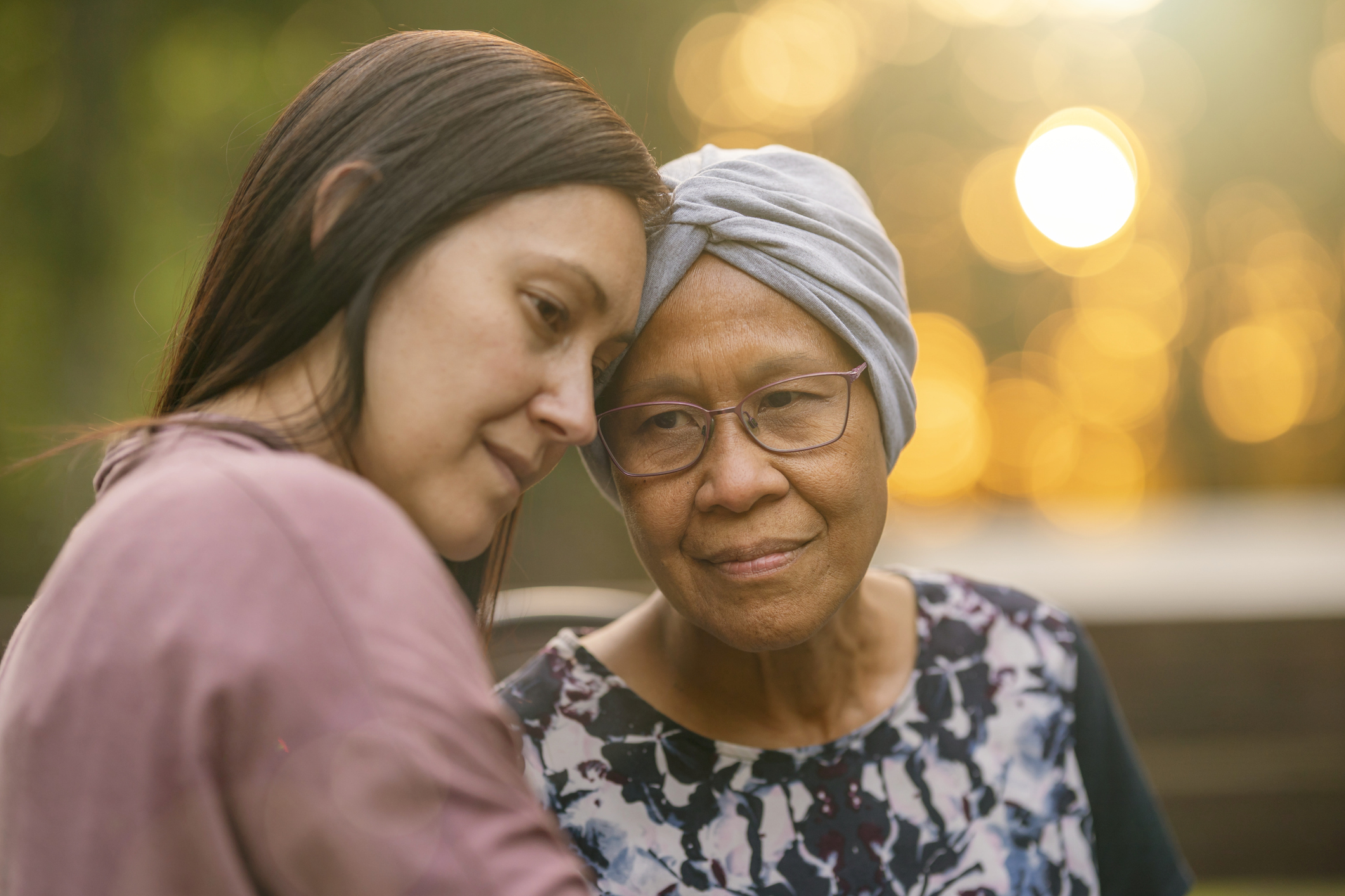 A senior woman with cancer smiles softly with a contemplative expression as her adult daughter embraces and comforts her.