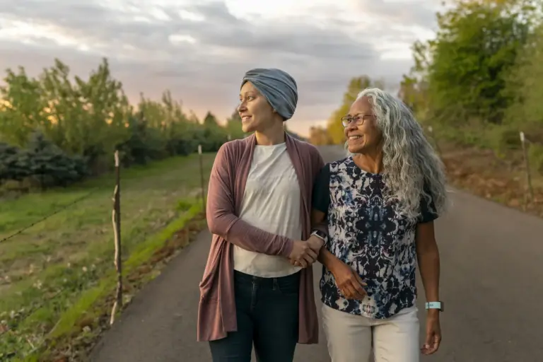 A young woman recovering from cancer walks arm in arm withcaregiver. They are enjoying time together while taking a relaxing walk down a rural road at sunset. Both women are smiling and looking off into the distance.