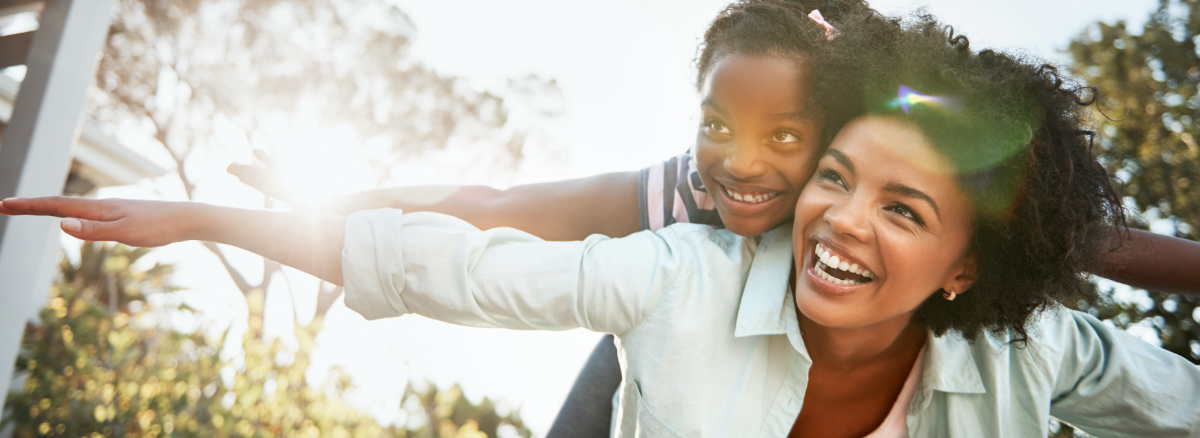 young girl and her mother smiling as her mother holds her on her back with their arms out like an airplane