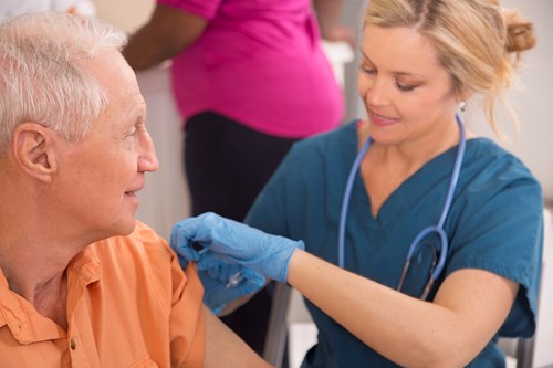 blonde nurse in blue scrubs giving senior male on orange shirt a flu shot