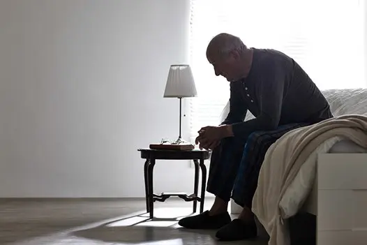 senior man sitting on unmade bed hands clasped head down